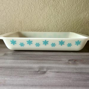 May include: Rectangular, off-white Pyrex baking dish with turquoise snowflake pattern. The dish has a slightly flared rim and is set against a neutral background. A vintage kitchenware item.