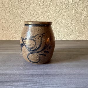 May include: A tan stoneware crock with a speckled texture and a dark blue hand-painted bird design on the front. The vessel features a rounded body, a short neck, and a thick rim, with a small set of initials marked near the bird illustration.