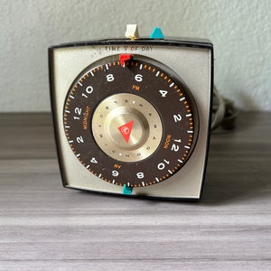 May include: A vintage electric timer with a brown and gold face, displaying the time of day. The face has a circular design with numbers and the words "MIDNIGHT" and "NOON". The timer has a silver frame and a cord.