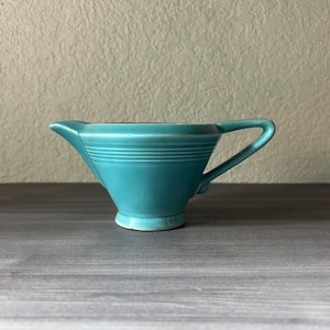 May include: A turquoise ceramic creamer with a handle and spout. The creamer has horizontal ridges around the body and a small footed base. The handle is curved and the spout is pointed.