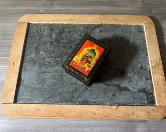May include: A small, rectangular black box with a colorful illustration of a person in a red and gold outfit. The box is on a slate chalkboard with a wooden frame.