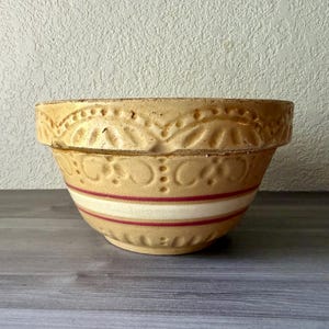 May include: A vintage beige ceramic mixing bowl with a decorative rim and red and white stripes. The bowl has a wide, rounded shape and is likely used for baking or food preparation. The bowl is sitting on a gray surface.