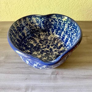 May include: A ceramic bowl featuring a deep blue and white speckled pattern throughout the interior and exterior. The bowl has a rounded shape with a distinct indentation along the top rim, creating a heart-like silhouette with a smooth, glossy finish.