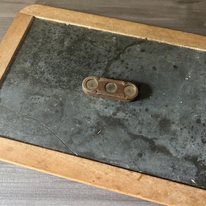 May include: An antique slate chalkboard with a wooden frame. A small, rectangular, brass-colored level tool with three circular viewing windows rests on the slate surface. The slate has a dark, mottled appearance.