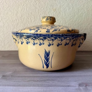 May include: A ceramic lidded casserole dish with a cream-colored base and blue floral and wheat stalk designs. The lid and rim feature blue floral patterns. The dish has two small handles and is suitable for serving or storage.