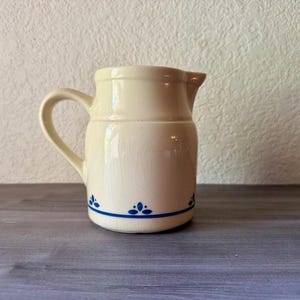 May include: Cream-colored ceramic pitcher with a curved handle and spout. The pitcher features a blue floral design along the base and a blue stripe. The pitcher is sitting on a gray wooden surface.