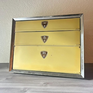 May include: A vintage kitchen organizer with three compartments, each labeled for foil, wax paper, and paper towels. The organizer has a gold-colored front with brown shield-shaped labels and a silver-colored metal frame.