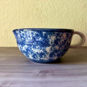 May include: A ceramic mixing bowl featuring a blue and white speckled pattern, a rounded body, a small pouring spout on the left side, and a single curved handle on the right side. The interior and exterior display a consistent mottled finish.