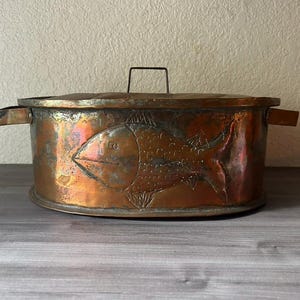 May include: A copper-colored metal container with a lid and handles. The container features an embossed fish design on the front. The lid has a small handle. The container is sitting on a wooden surface.