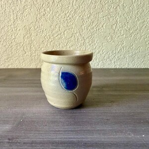 May include: A small, round, beige stoneware crock featuring a smooth, matte finish and a single, vibrant cobalt blue leaf-shaped accent painted on the front. The vessel has a thick, slightly flared rim and a sturdy, rounded base design.