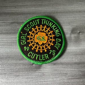 May include: A circular Girl Scout Thinking Day patch from 1975. The patch features a black background with green and gold embroidered details. The text "GIRL SCOUT THINKING DAY" and "CUTLER" are visible.