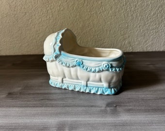 May include: A white ceramic baby cradle planter with blue accents. The cradle features a curved hood, ruffled trim, and decorative blue roses. The planter is designed to hold small plants or flowers, adding a touch of vintage charm.