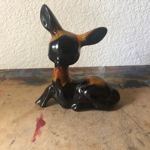 May include: A ceramic deer figurine in a resting pose. The deer is primarily black with accents of orange and brown on the neck and back. The word "Niagara" is written on the side. The figurine has a glossy finish.