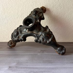 May include: Ornate, dark brown metal stand with a central circular opening and three decorative legs. The stand features detailed carvings and a weathered finish, suggesting an antique or vintage aesthetic. The legs have rounded feet.