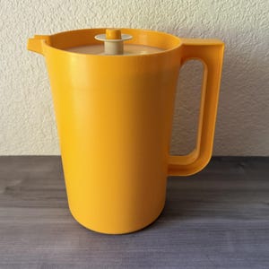 May include: A bright yellow plastic pitcher featuring a tall cylindrical body, a sturdy side handle, and a pouring spout. The pitcher includes a matching yellow lid with a small, light beige circular knob centered on top for easy removal.