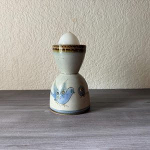 May include: A ceramic egg cup with a blue bird design. The egg cup is light beige with brown accents and a blue bird painted on the side. A white egg sits in the top of the cup. The cup is on a gray surface.