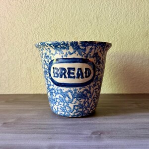 May include: A ceramic bread crock featuring a blue and white sponge-painted or splatter pattern. The container has a flared rim and a central oval-shaped plaque with the word BREAD embossed in dark blue lettering against a white background.