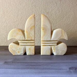 May include: Two matching bookends in a fleur-de-lis design. The bookends are carved from a creamy, off-white stone with subtle tan veining. Each bookend has a rounded base and a vertical, stylized floral shape.