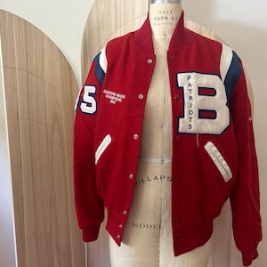 May include: A red varsity jacket with white and blue accents. The jacket features the word "Patriots" and the letter "B" on the front, along with the number "15" and text that reads "Eastern Mass Champions 2007".
