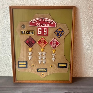 May include: Framed vintage Cub Scout memorabilia. The display features a tan felt backing with various patches and badges, including the text "SOUTH FLORIDA COUNCIL" and the number "69". The frame is a warm brown color.