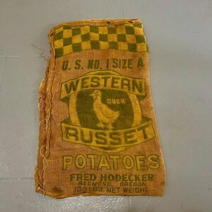 May include: Vintage burlap potato sack with a green and yellow checkered top. The sack features the text "U.S. No. 1 Size A Western Duck Russet Potatoes" with a duck graphic. The sack reads "Fred Hodecker, Redmond, Oregon, 100 lbs Net Weight."