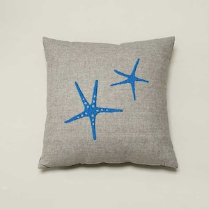 May include: A gray pillow with two blue embroidered starfish. The pillow is made of a soft, textured fabric.