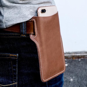 May include: A brown leather phone holster with white stitching. The holster is attached to a belt and has a phone inside.