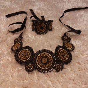 Può includere: Un set di collana e braccialetto nero e oro con un design circolare. I gioielli sono realizzati in tessuto e hanno un bordo di perline.