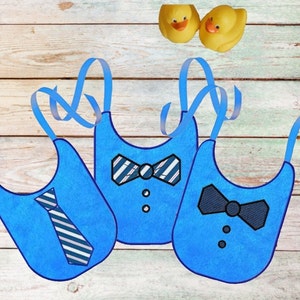 May include: Three blue baby bibs with different embroidered designs: a tie, a striped bow tie, and a solid bow tie. Two yellow rubber duckies are in the background. The bibs have blue straps.