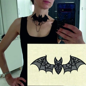 Puede incluir: Un collar gargantilla de encaje negro en forma de murciélago con un colgante de cristal. El collar está puesto en el cuello de una persona. Una imagen separada muestra un murciélago negro con intrincados detalles de encaje sobre un fondo de color crema.