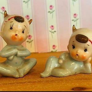 May include: Two ceramic figurines of infants wearing light blue onesies with small gold horns on their heads. One infant sits cross-legged with hands together, while the other reclines on one side. Both have painted facial features and dark hair.