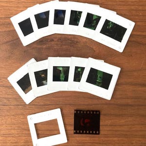 May include: A collection of white plastic slide mounts with a variety of images inside. There is also a single roll of 35mm film.