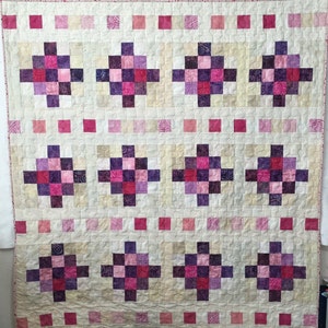 May include: A quilt with a patchwork design featuring a repeating pattern of squares in shades of pink, purple, and white. The quilt is edged with a pink border.