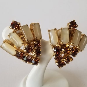May include: A pair of gold tone clip-on earrings featuring brown and white stones. The earrings are shaped like a fan and have a geometric design.