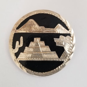 May include: A silver and black circular pin with an image of a pyramid, mountains, and a cactus. The pin is a traditional Mexican design.