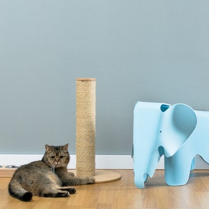 May include: A brown tabby cat lies on a wooden floor next to a scratching post and a blue elephant-shaped stool.