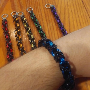 May include: A collection of chainmail bracelets featuring interlocking metal rings. One bracelet is blue and black, while the others feature black rings accented with red, orange, yellow, green, or purple rings, each finished with a silver-tone ring clasp.