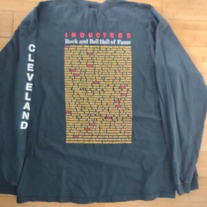 May include: A dark green long-sleeve t-shirt with white text on the sleeve that says "CLEVELAND". The back of the shirt has white text that says "INDUCTEES Rock and Roll Hall of Fame" and a list of inductees in yellow and red text.