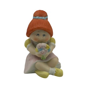 May include: A porcelain figurine of a little girl with red hair, wearing a pink dress and holding a bouquet of flowers.