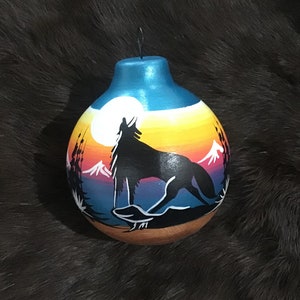 May include: Hand-painted gourd ornament featuring a howling wolf silhouette against a sunset backdrop. The design includes a full moon, mountains, and trees. The ornament has a blue top and a wire for hanging.