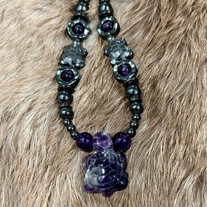 May include: A handcrafted necklace featuring a dark gray beaded chain with amethyst and turtle-shaped pendants. The necklace has a central turtle pendant carved from amethyst, with smaller turtle and flower-shaped accents.