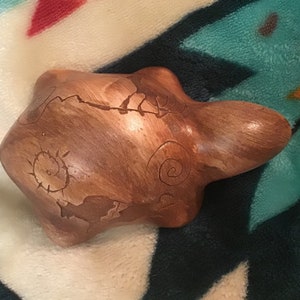 May include: A wooden turtle sculpture with intricate carvings of a sun, a spiral, and other designs. The turtle is brown and has a smooth, polished finish.