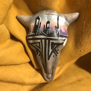 May include: A hand-painted ceramic bull skull with a southwestern design. The skull is painted in shades of gray, black, purple, and pink. The design features a stylized landscape with mountains and a sunset.