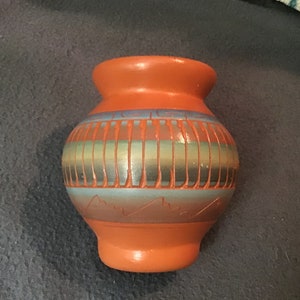 Pode incluir: Um pequeno pote de barro laranja com um desenho geométrico em tons de azul, verde e marrom. O pote tem um corpo arredondado e uma borda ligeiramente alargada.