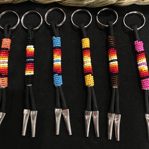 May include: Six keychains with beaded accents in various colors, including orange, black, red, yellow, blue, and pink. Each keychain has a black cord and a silver-colored metal fork-like charm. The keychains are arranged on a dark surface.