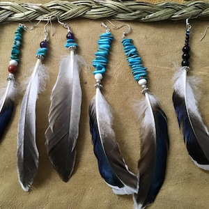 May include: A collection of six long dangle earrings featuring natural feathers with white, grey, and dark blue tips. The earrings are accented with various beads including turquoise chips, round white, brown, purple, and black beads on silver hooks.