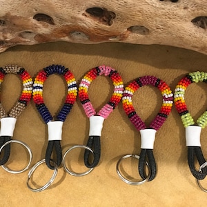 Op de afbeelding: Vijf ronde sleutelhangers met gedetailleerd kraalwerk in diverse kleurpatronen, waaronder beige, blauw, roze, magenta en limoengroen. Elke sleutelhanger heeft een witte band die de kralenlus verbindt met een zwart koord en een zilverkleurige metalen ring.