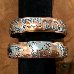 Peut inclure: Deux bracelets en métal cuivré et argenté. Le bracelet supérieur présente des motifs gravés de formes géométriques et d'un oiseau stylisé. Le bracelet inférieur a des chevaux gravés. Les bracelets sont exposés sur un support noir.