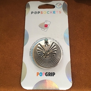 May include: A silver PopSockets PopGrip with a circular design featuring a maze and a small figure. The PopGrip is attached to a white backing card with the PopSockets logo and product information.