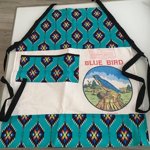 Puede incluir: Delantal turquesa con un patrón geométrico y un bolsillo blanco. El delantal presenta un gran gráfico de un pájaro azul con el texto "BLUE BIRD". Lazos y ribetes negros completan el diseño. El delantal es probablemente para cocinar o hacer manualidades.